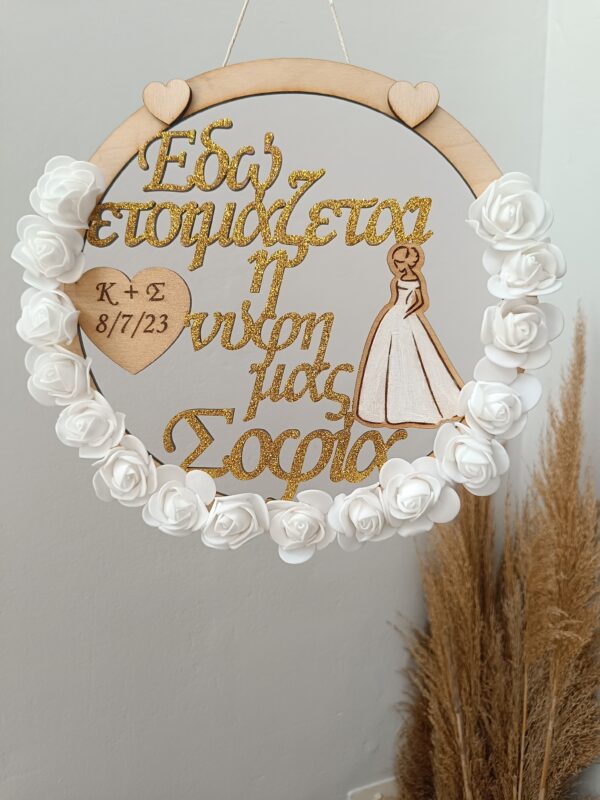 Χειροποίητο ξύλινο διακοσμητικό στεφάνι “Bride” στολισμένο με λευκά τριαντάφυλλα και χρυσόσκονη. Χαραγμένα τα αρχικά του ζευγαριού και η ημερομηνία του γάμου.