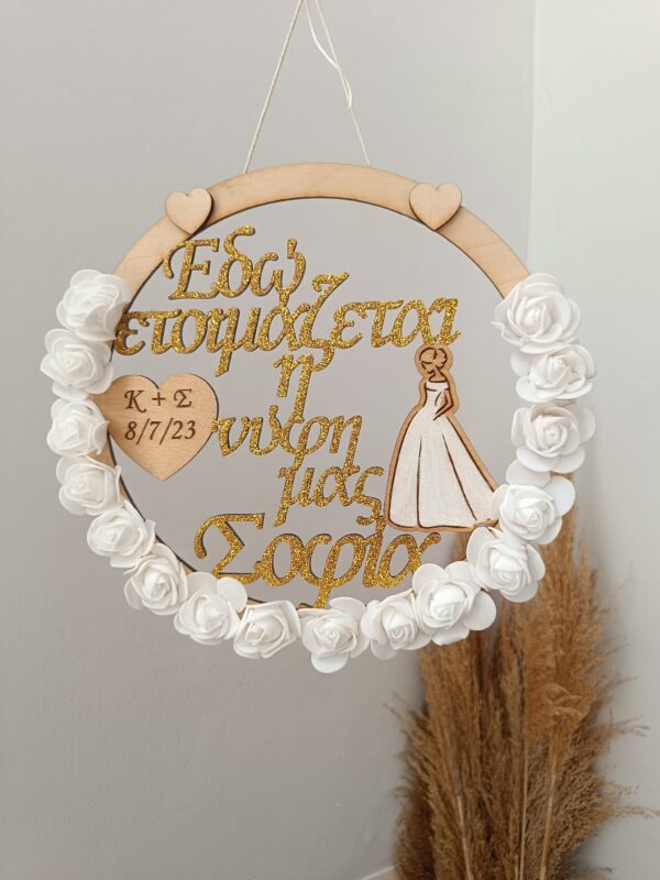 Χειροποίητο ξύλινο διακοσμητικό στεφάνι “Bride” στολισμένο με λευκά τριαντάφυλλα και χρυσόσκονη. Χαραγμένα τα αρχικά του ζευγαριού και η ημερομηνία του γάμου.